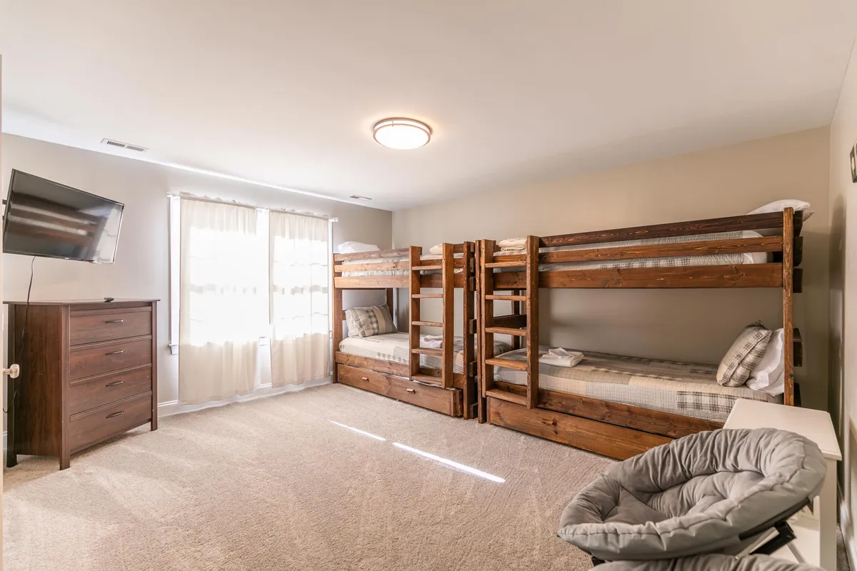 A spacious bunk room designed with multiple built-in bunk beds, a window dressed with curtains, a dresser, a television, and a comfortable chair. The ambiance is friendly and welcoming, perfectly suited for children or teenagers to sleep, play, and create shared memories.