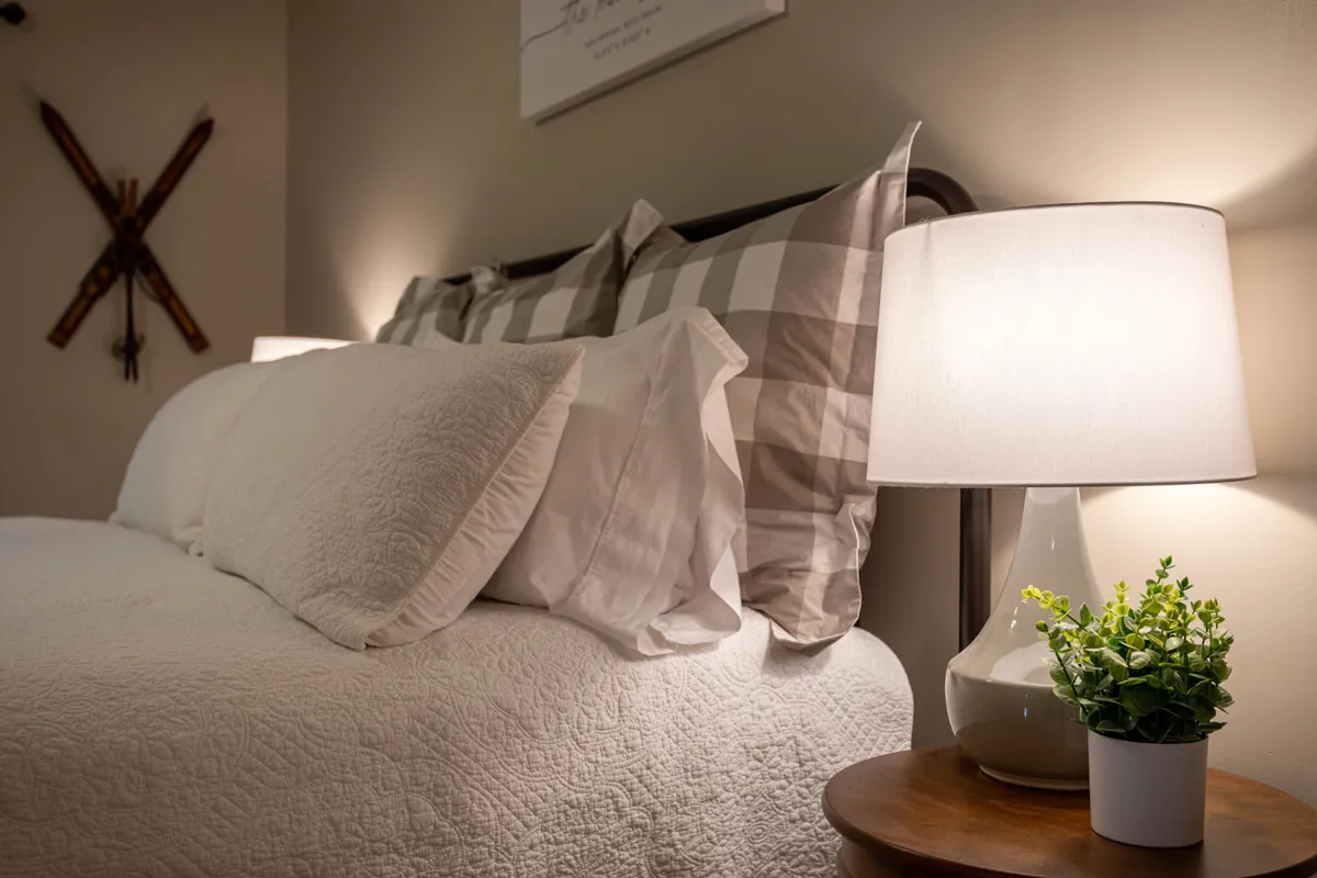A cozy guest bedroom with a bed dressed in decorative pillows, a nightstand topped with a lamp and a small plant, and subtle wall art. The space feels tranquil and comforting, ideal for unwinding and enjoying peaceful moments.