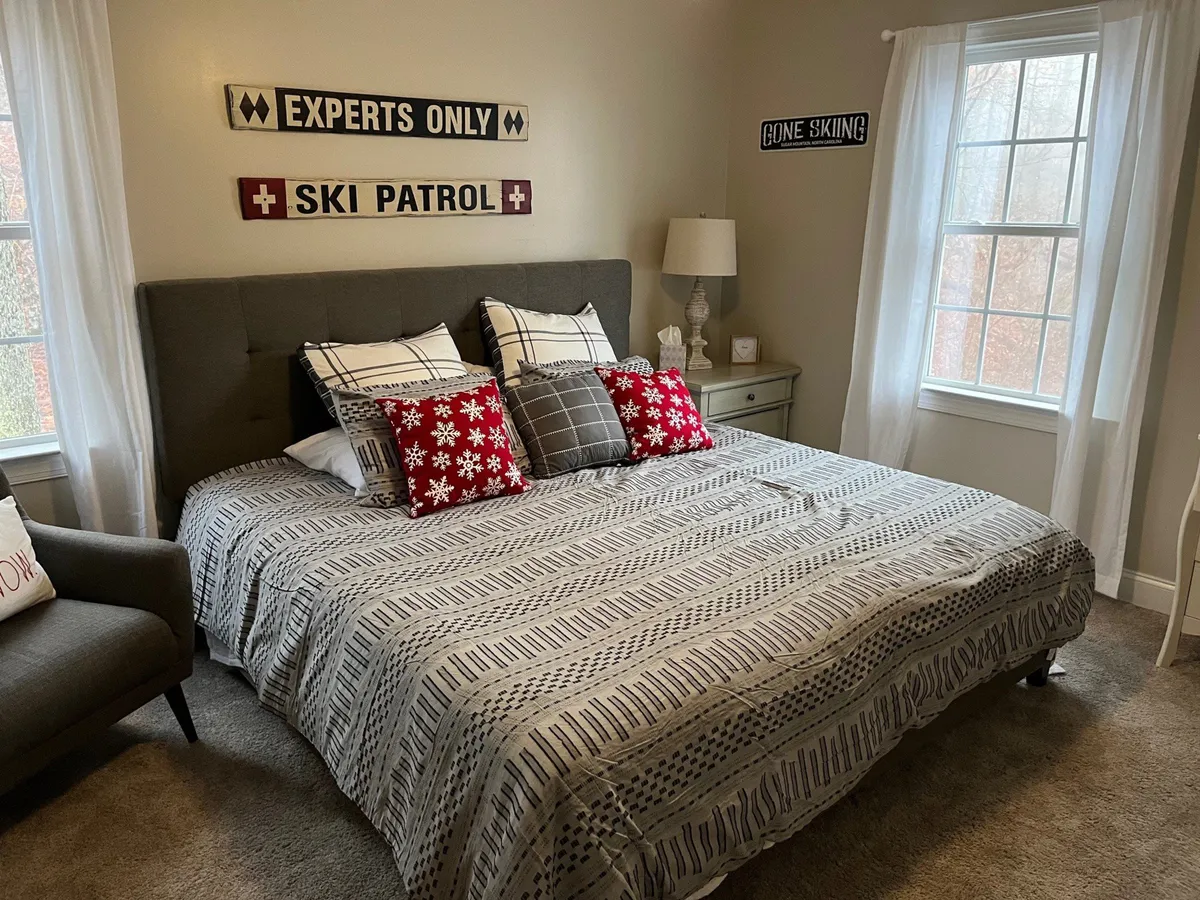 A bedroom with patterned bedding, decorative throw pillows, and wall signs, evoking warmth and comfort. The window provides a picturesque view, reinforcing the sense of connection to the outdoors. The overall setting feels private and tranquil, perfect for unwinding together.