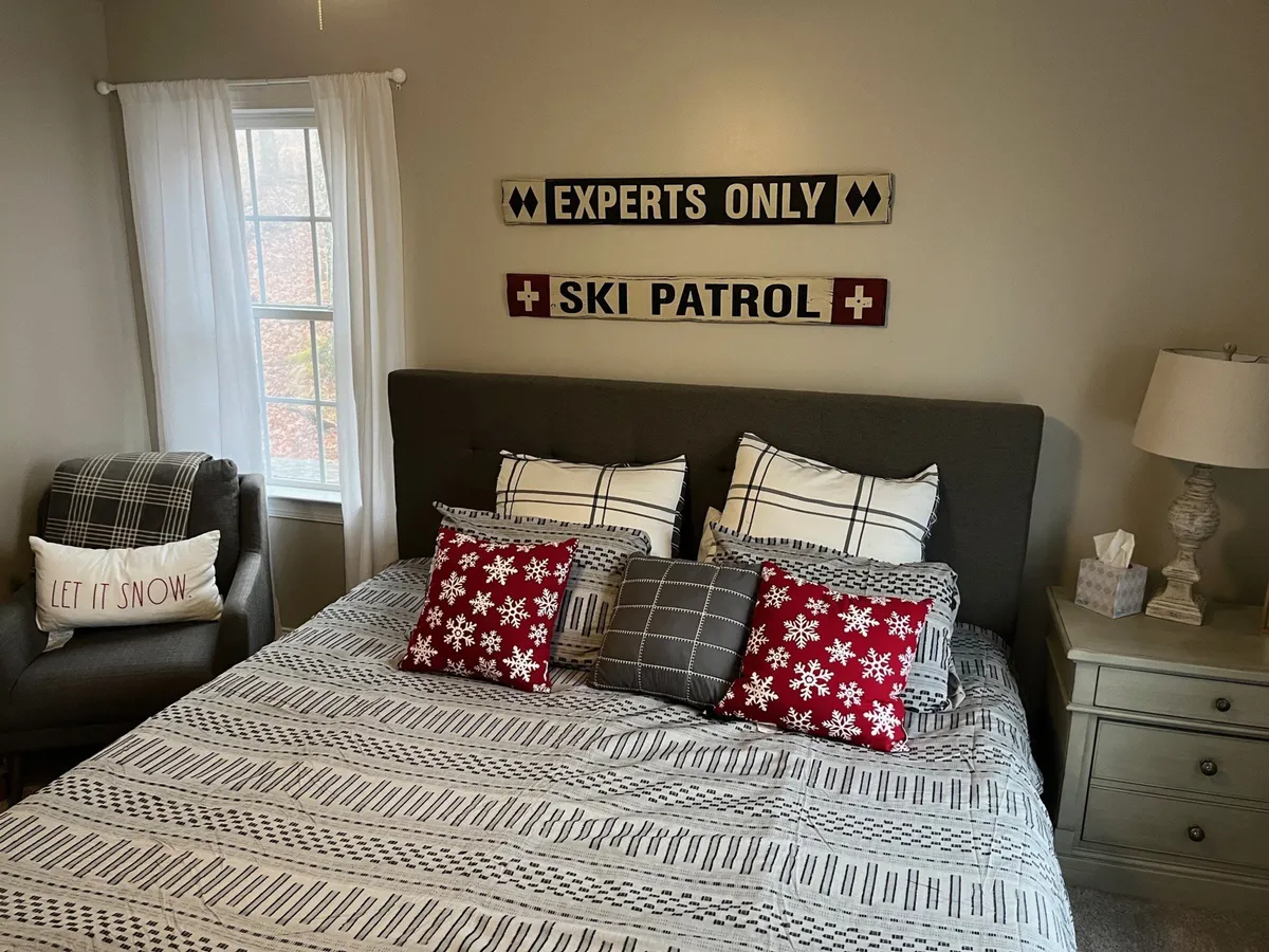 A warmly styled bedroom featuring a bed with decorative pillows, ski-themed wall decor, and a window letting in abundant natural light. The ambiance is cozy and welcoming, with hints of mountain charm visible in the decor and the inviting glow from outside.
