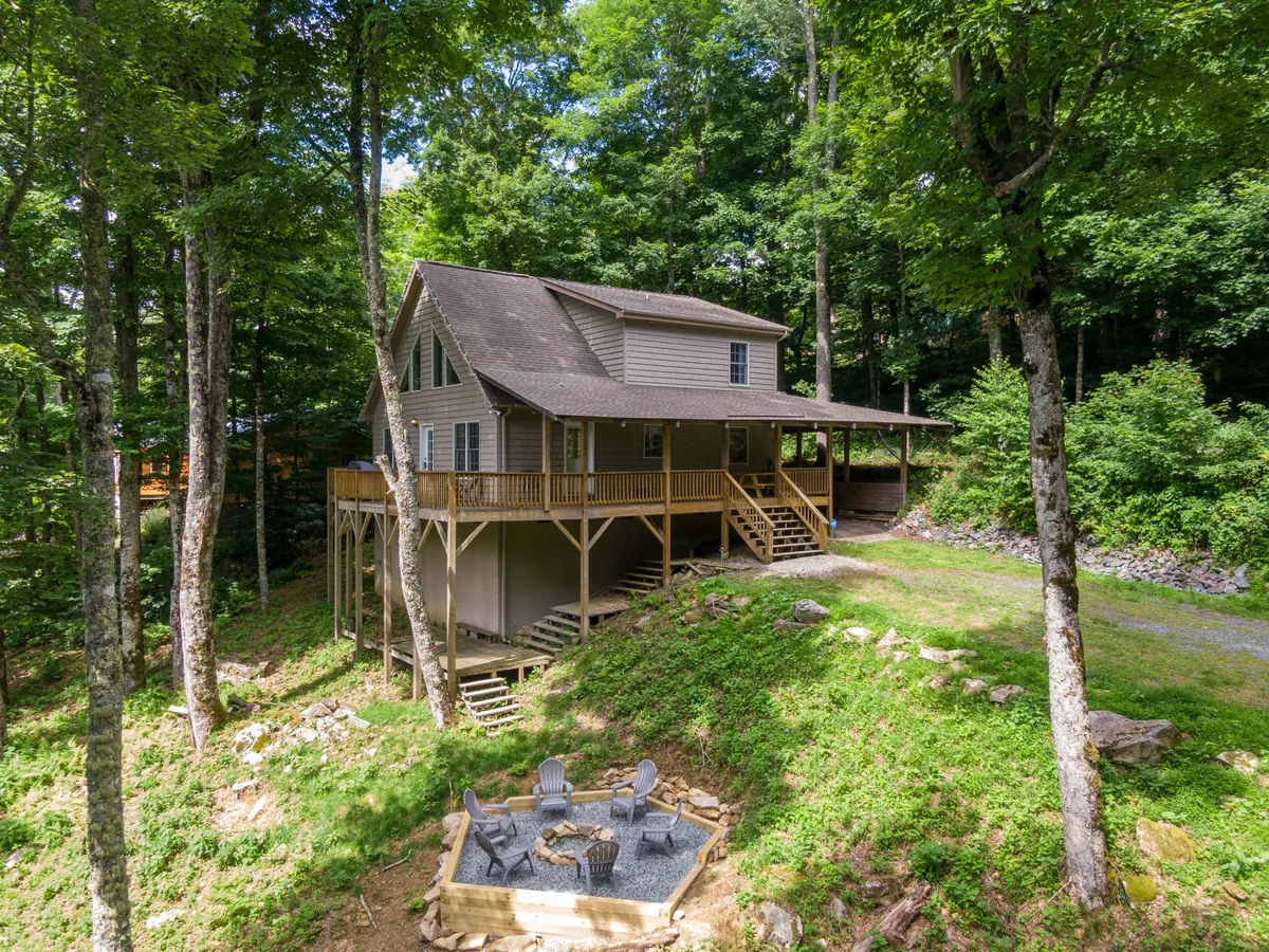 The exterior view showcases the house nestled among tall trees with a cozy fire pit area in the foreground. The inviting outdoor space encourages gatherings and stargazing after a day on the slopes.