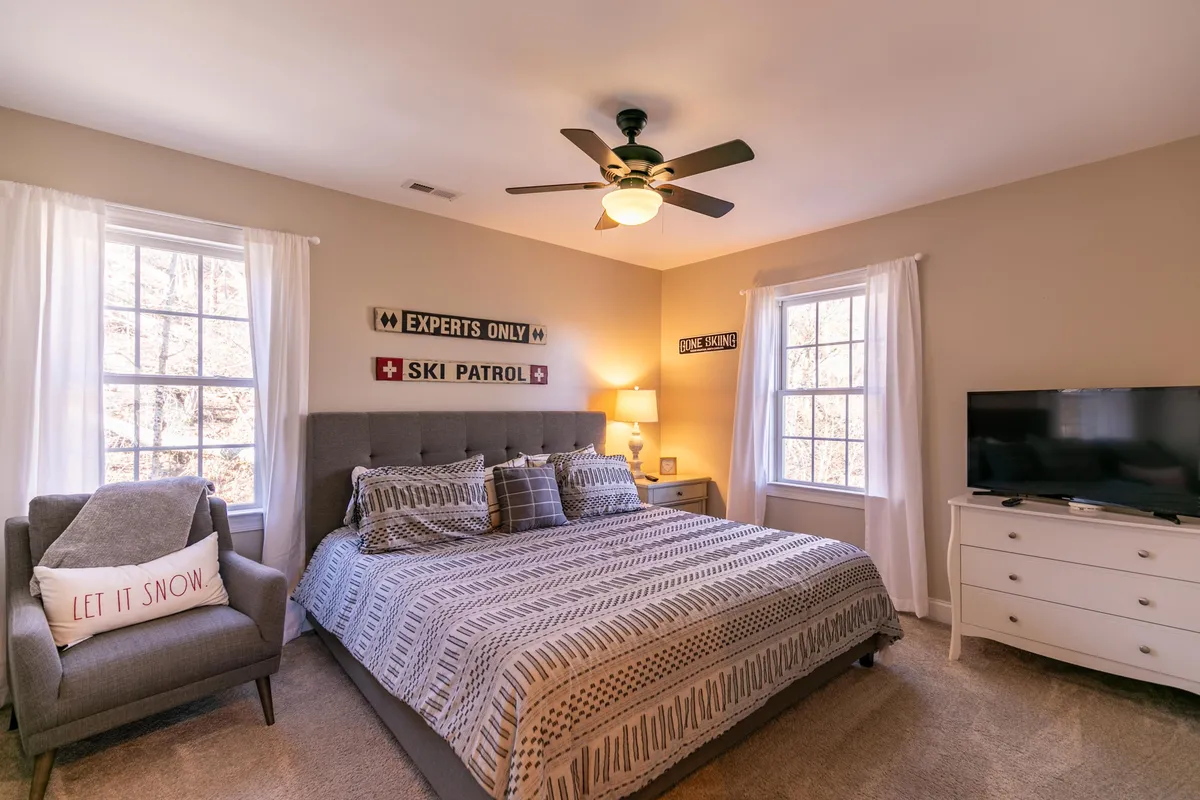 A welcoming bedroom with patterned bedding, an armchair adorned with a decorative pillow, and expansive windows that flood the room with natural light. Wall signs add a homely touch, creating an atmosphere perfect for relaxing or sharing stories after a day of adventure.