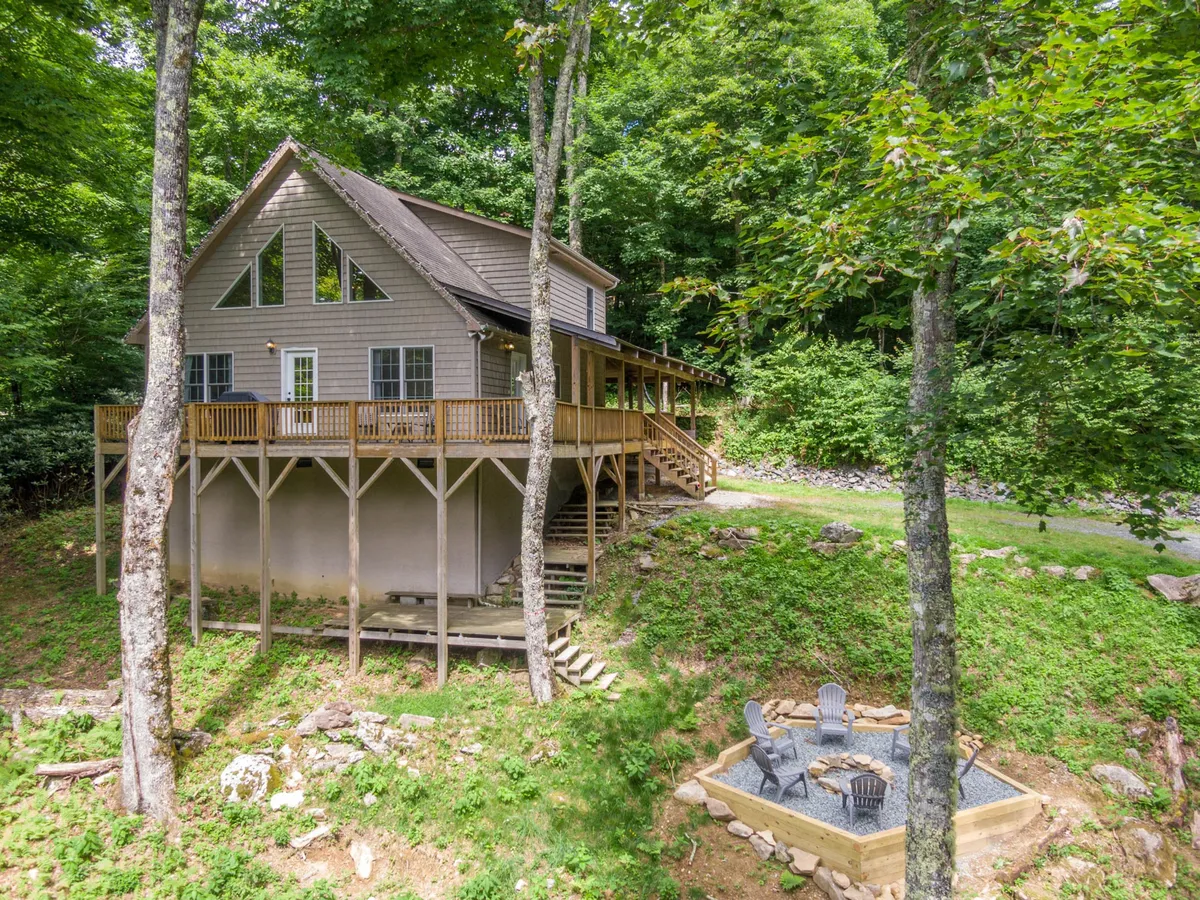The hero image showcases a charming single-family house nestled among tall trees, featuring a spacious deck perfect for sunset views and a cozy fire pit area surrounded by Adirondack chairs. The serene setting highlights the inviting outdoor spaces, with hints of mountain wilderness and a sense of privacy, making it ideal for gatherings and relaxation after days spent exploring nearby ski slopes or town attractions.