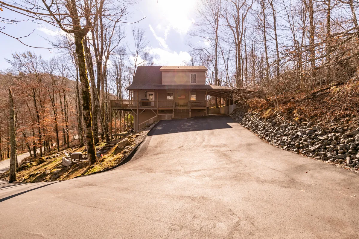 A broad view of the house with its welcoming porch, newly paved driveway, and the surrounding woods. The serene and secluded environment is clearly visible, suggesting privacy and easy access year-round.