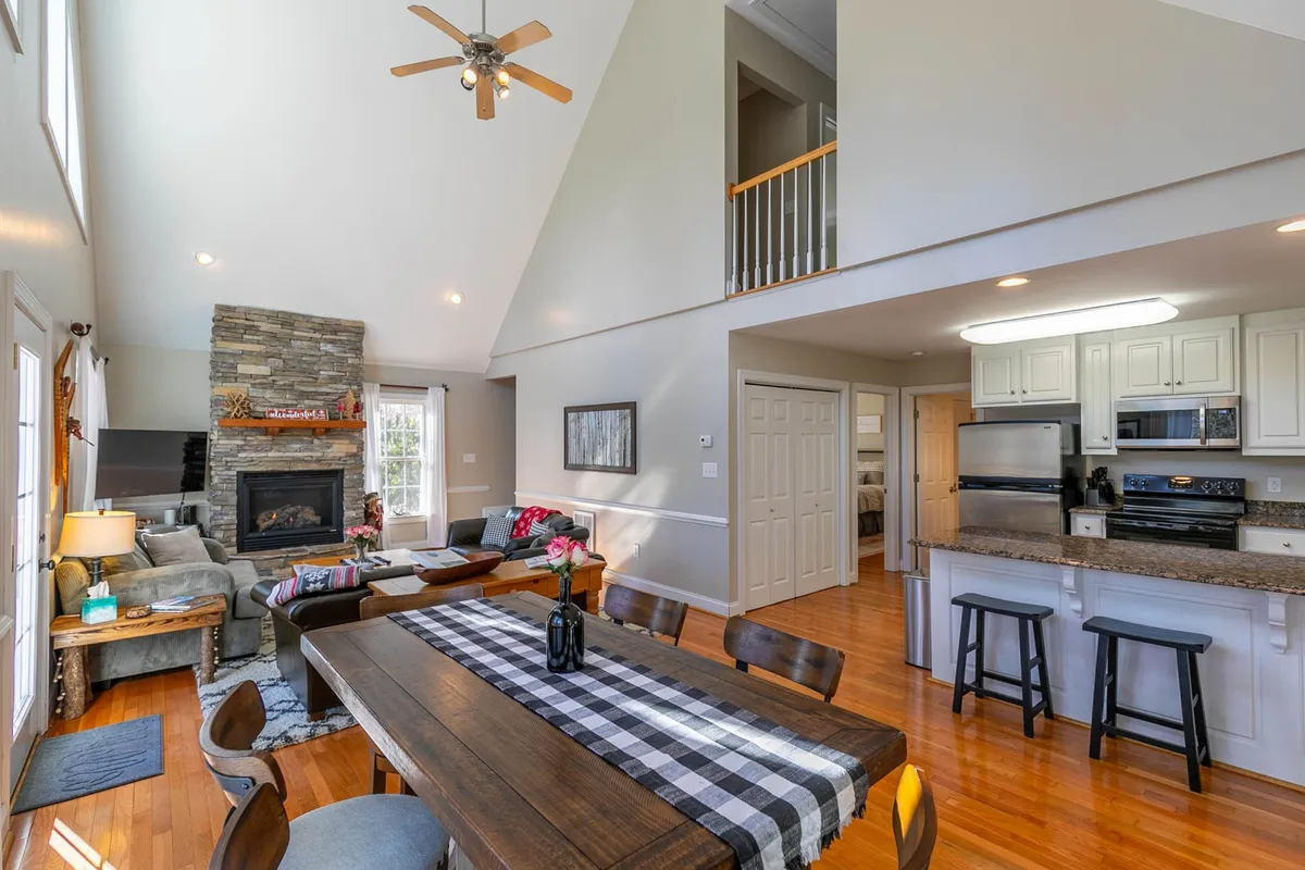 This interior shot displays an open-plan space featuring a stone fireplace, a dining table, and a visible open kitchen. The arrangement highlights the seamless flow between the living, dining, and kitchen areas. Large windows fill the room with natural light, while cozy furniture beckons guests to gather and relax.