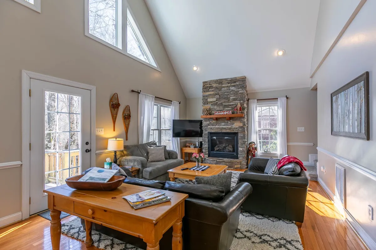 A cozy living room with a striking stone fireplace, large bright windows, and plush seating. The space is styled for comfort, ideal for gatherings, quiet moments, or winding down after a day outdoors. The blend of light and warmth suggests relaxation and security.